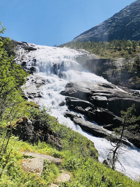 A wide waterfall cascades down rocky cliffs surrounded by green trees and vegetation under a clear blue sky.