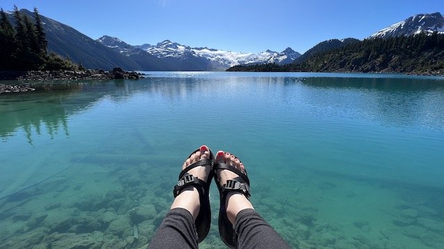 A person wearing sandals sits by a clear blue lake, with snow-capped mountains visible in the distance under a clear sky.