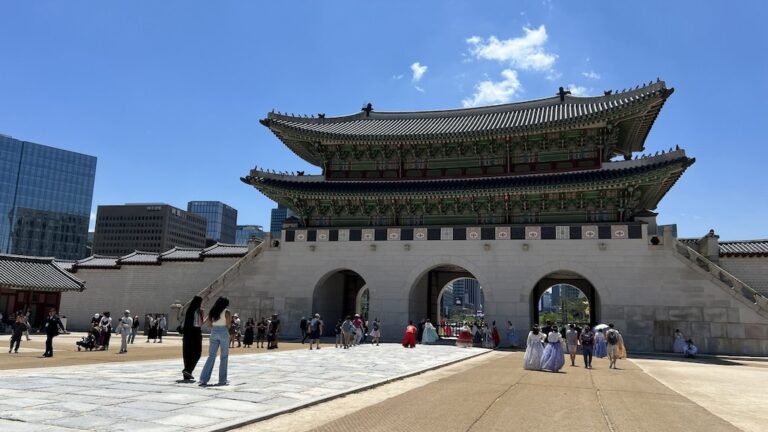 People walk near the main gate of a traditional Korean palace, with modern buildings in the background under a clear blue sky.
