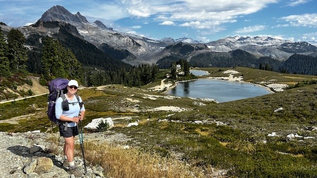 A hiker with a backpack stands on a trail overlooking a lake and mountains under a partly cloudy sky.