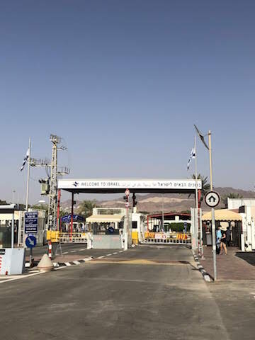 Border crossing checkpoint with a sign reading Welcome to Israel under a clear sky, with gates, barriers, and Israeli flags visible.