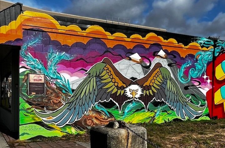 Colorful mural of a bird with outstretched wings, mountains, clouds, and abstract patterns painted on a building wall.