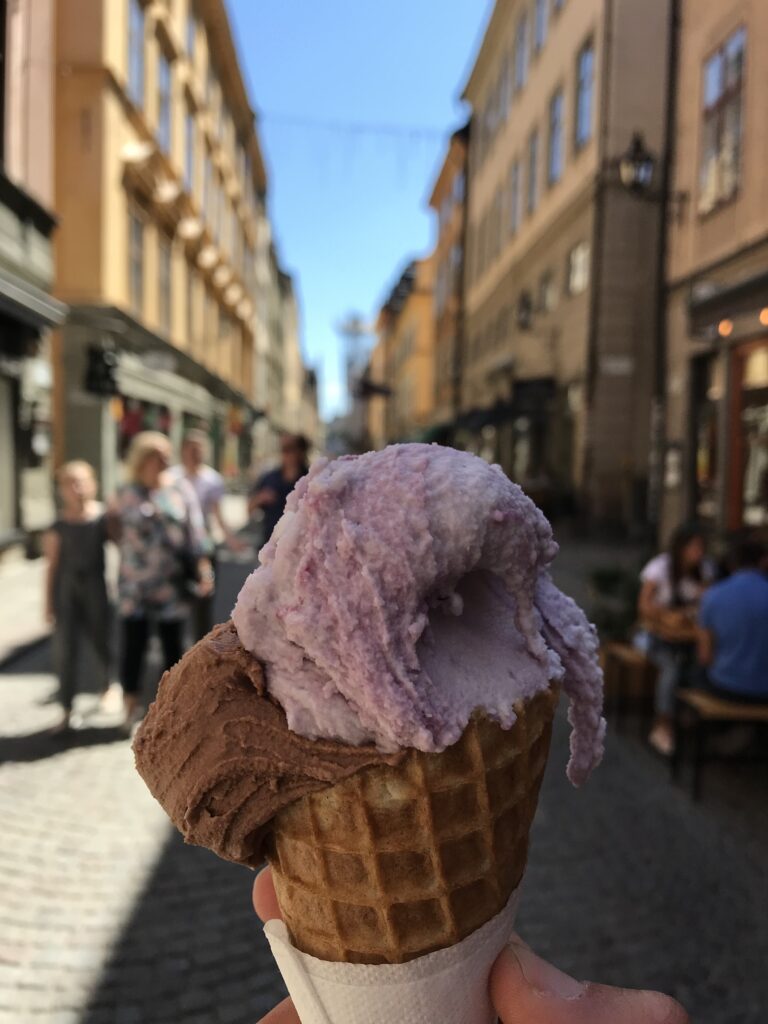 A hand holds an ice cream cone with two scoops on a cobblestone street lined with buildings and people walking in the background.