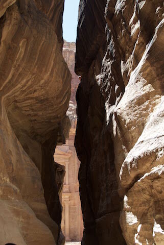 Tall sandstone cliffs with a narrow passage reveal a glimpse of an ancient building facade in the background.