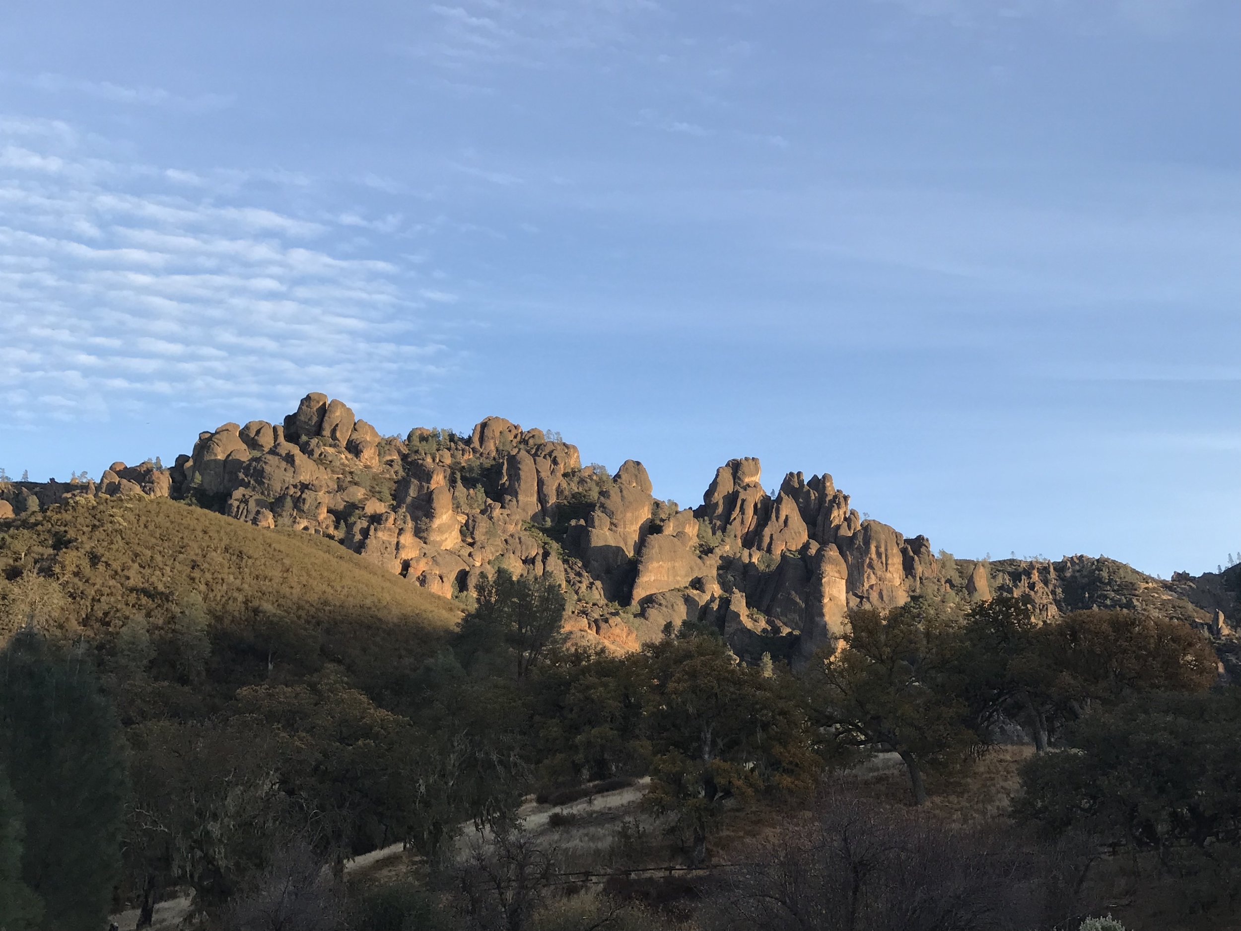 Rocky hill formation with scattered trees and bushes under a partly cloudy sky at sunset.