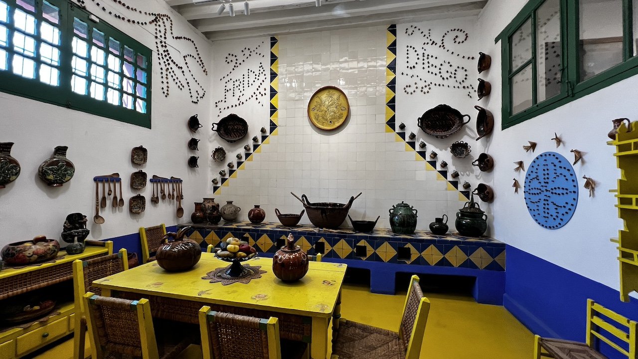 A kitchen with yellow furniture, decorative pottery, and colorful tiles on the walls and counters, with windows on either side.