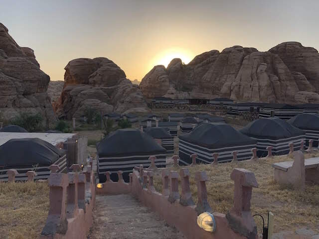 A row of black-and-white striped tents is set among rocky desert cliffs at sunset, with a stone walkway in the foreground.