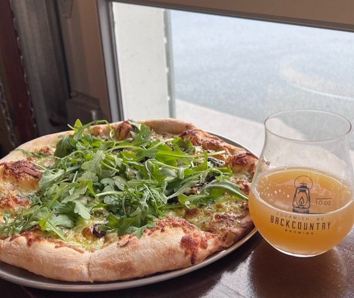 A pizza topped with arugula sits on a plate next to a glass of beer on a wooden table by a window.