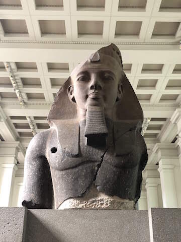 A large stone bust of an ancient Egyptian pharaoh is displayed indoors, showing visible cracks and damage.