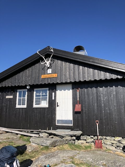 A black wooden mountain cabin with antlers above the door, two windows, and red shovels leaning against the wall under a clear blue sky.