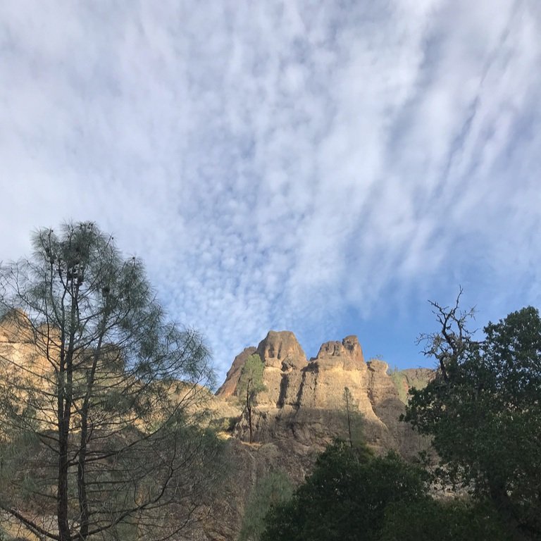 Rocky cliffs rise above trees under a blue sky with scattered, thin clouds.