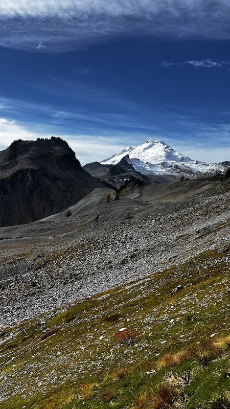 Snow-capped mountain peak under a blue sky with rocky and grassy terrain in the foreground.