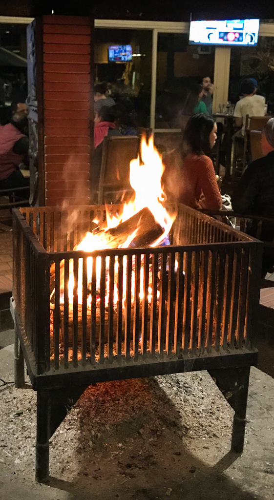 A fire burns in a metal grill at an outdoor seating area, with people sitting at tables in the background.