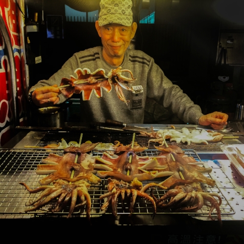 A street vendor grilling and displaying skewered squid at a food stall.