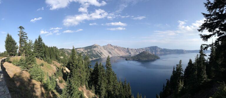 Panoramic view of a deep blue crater lake surrounded by forested cliffs and distant mountains under a partly cloudy sky.
