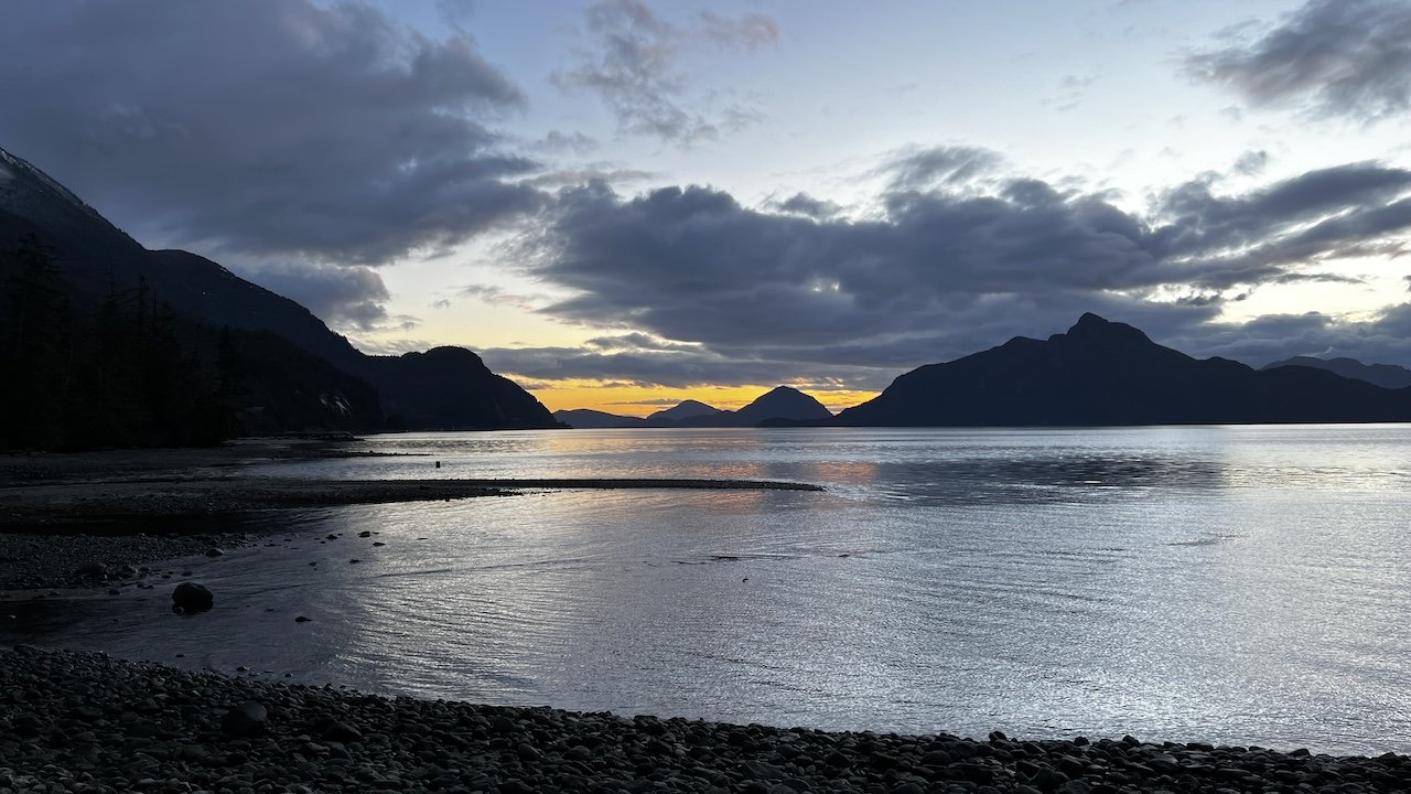 Calm coastal waters at sunset with mountains and clouds in the background, and a rocky shoreline in the foreground.