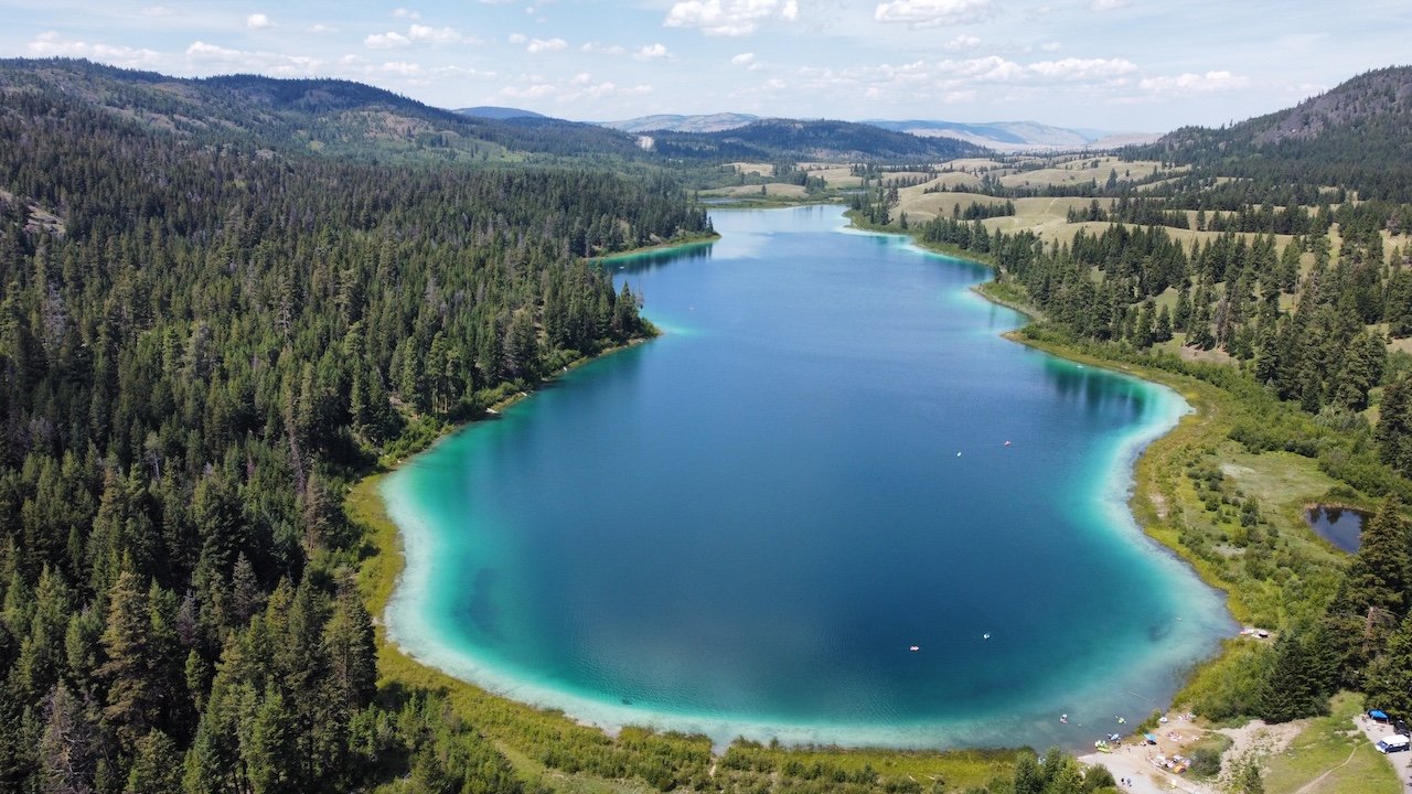 A turquoise lake surrounded by dense forests and hills under a partly cloudy sky.
