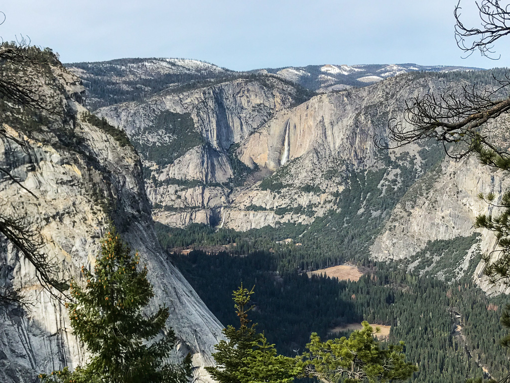 View of rugged granite cliffs and forested valley in Yosemite National Park, with sparse trees in the foreground and mountains in the background.