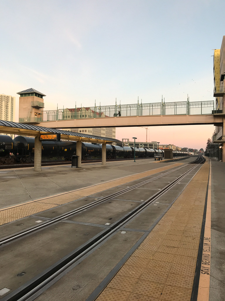 A train station platform with railroad tracks, a pedestrian bridge overhead, and trains parked alongside.