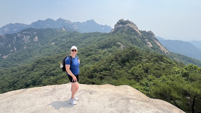 Person in athletic wear stands on a rocky outcrop overlooking forested mountains under a hazy sky.