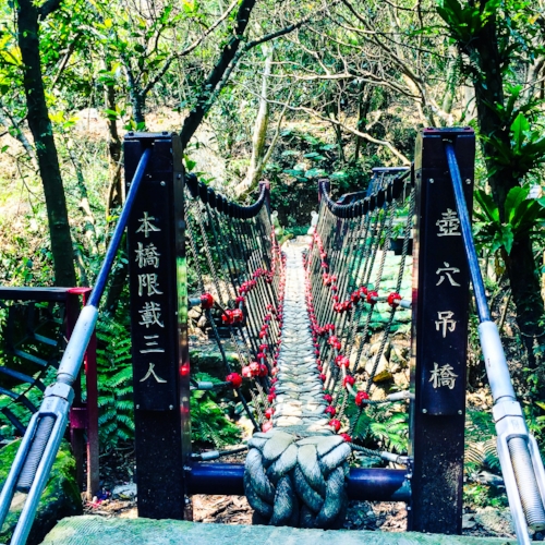 A narrow rope bridge crosses a rocky stream in a lush forest, with signs displaying Asian script on either side of the entrance.