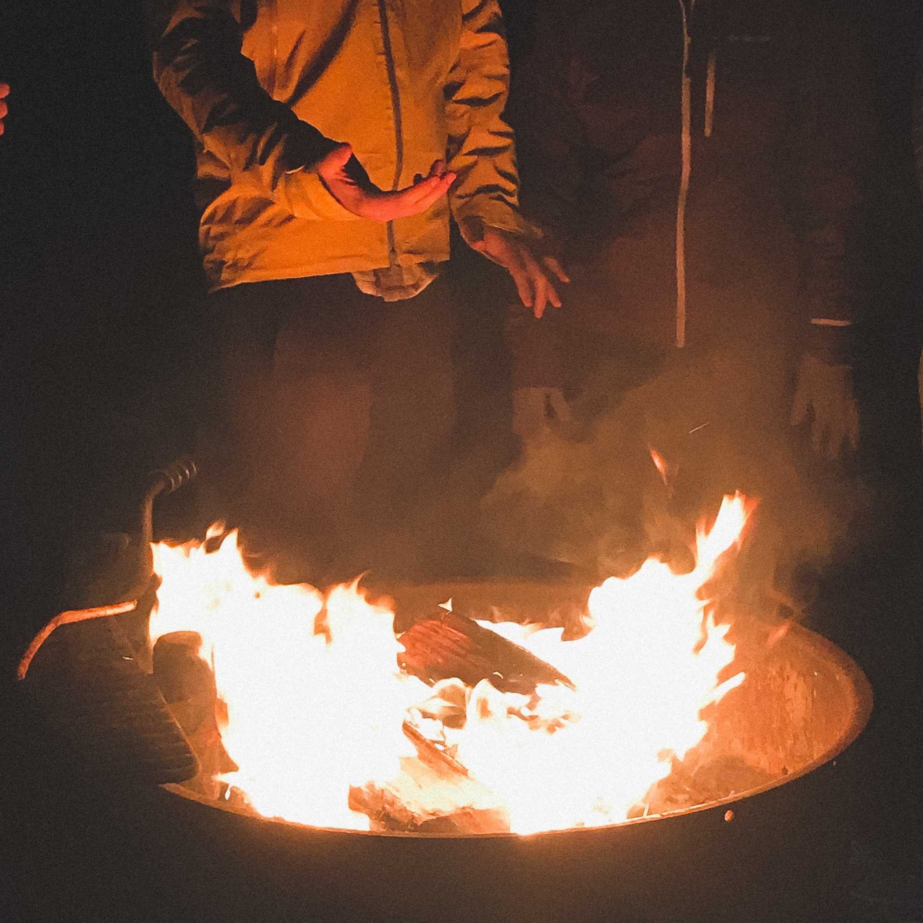 A person standing behind a raging fire in a pit at a campsite.