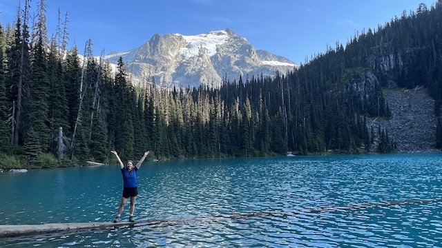 Person stands on a log in a blue lake surrounded by evergreen trees, with a snow-capped mountain in the background under a clear sky.
