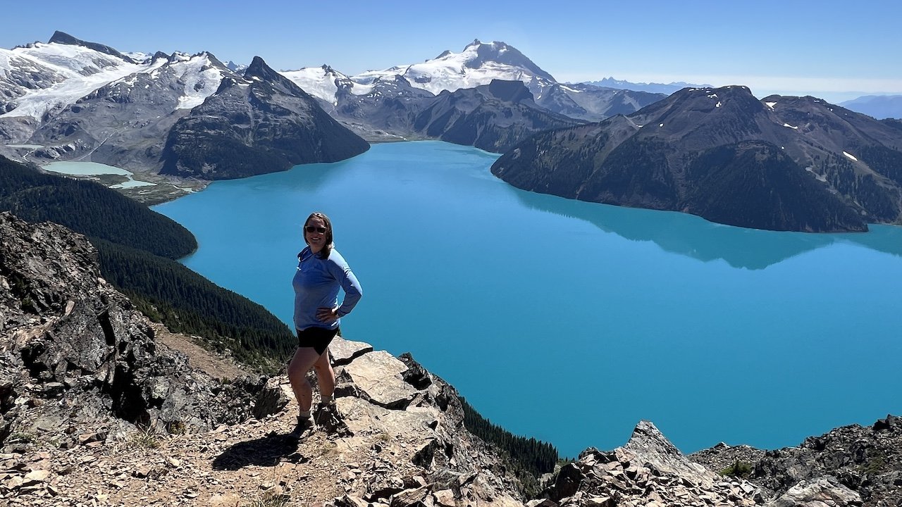 A person stands on a rocky mountain trail overlooking a bright blue lake surrounded by snow-capped peaks under a clear sky.