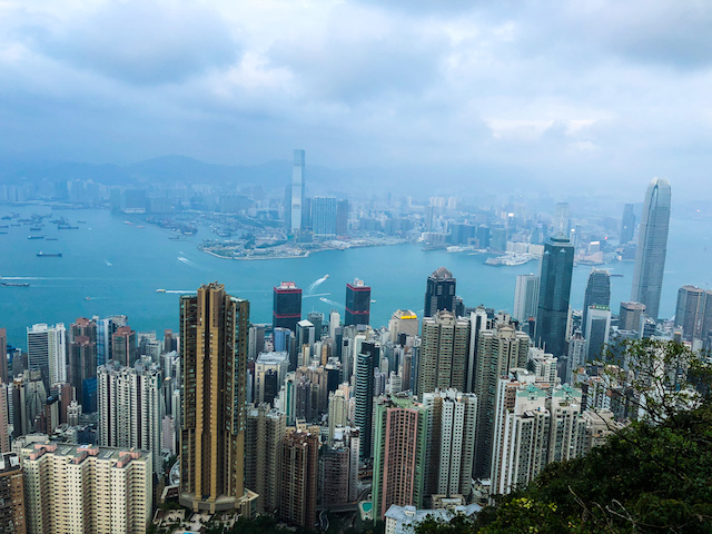 Aerial view of Hong Kong’s skyline with tall buildings, Victoria Harbour, and distant mountains under a cloudy sky.