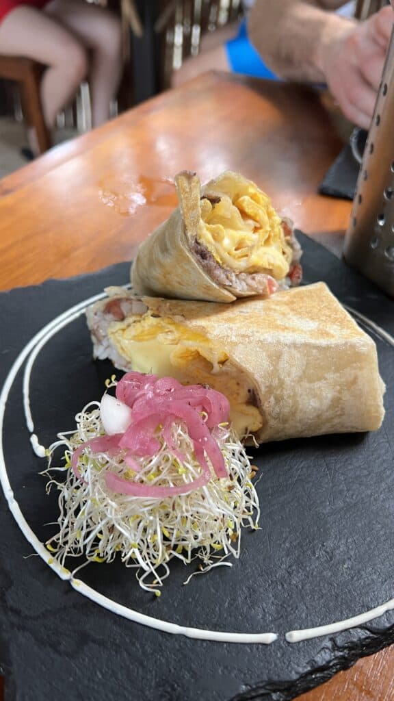 A breakfast burrito cut in half on a slate plate, served with alfalfa sprouts and pickled onions—perfect for your Tulum travel mornings.