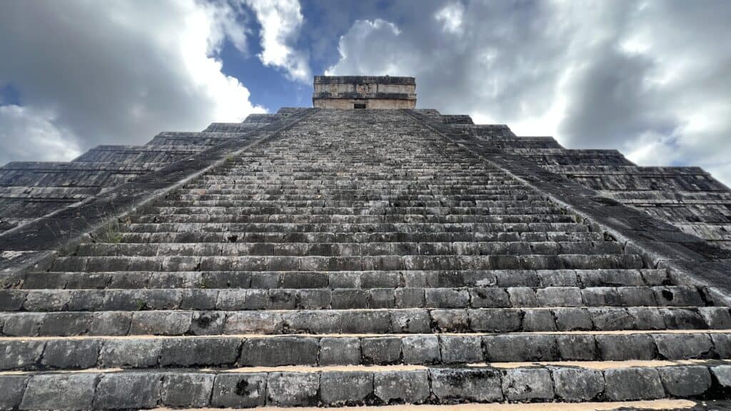 Stone steps lead up to the top of a large ancient pyramid under a partly cloudy sky—a must-see on your Tulum Travel adventure.