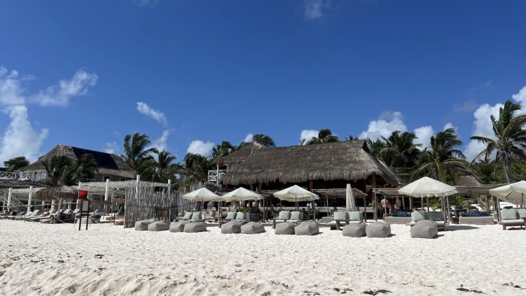 A Tulum beach resort with lounge chairs, umbrellas, and a thatched-roof building set among palm trees and a clear blue sky.