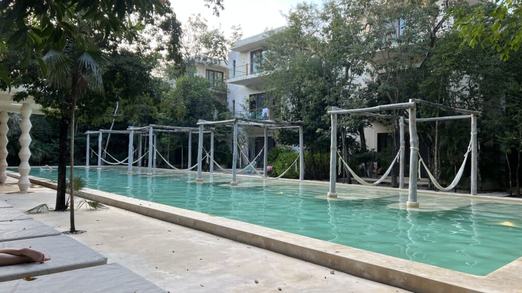A long outdoor pool in Tulum with hammocks over the water, surrounded by trees and modern white buildings—your Tulum Travel guide to relaxation.