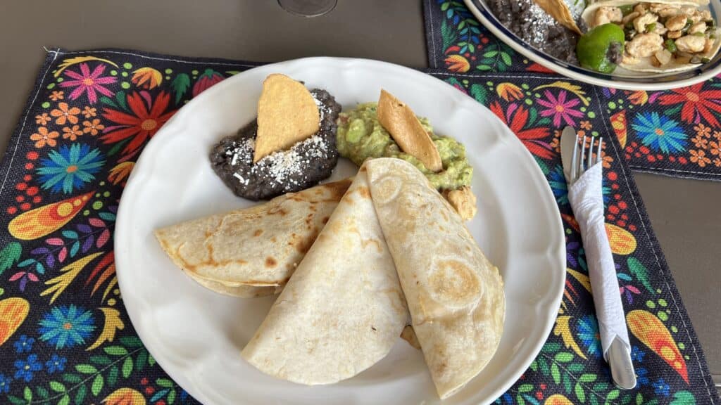 A plate with two folded tortillas, refried black beans with cheese, guacamole, and chips on a colorful placemat—perfect for your Tulum travel guide.