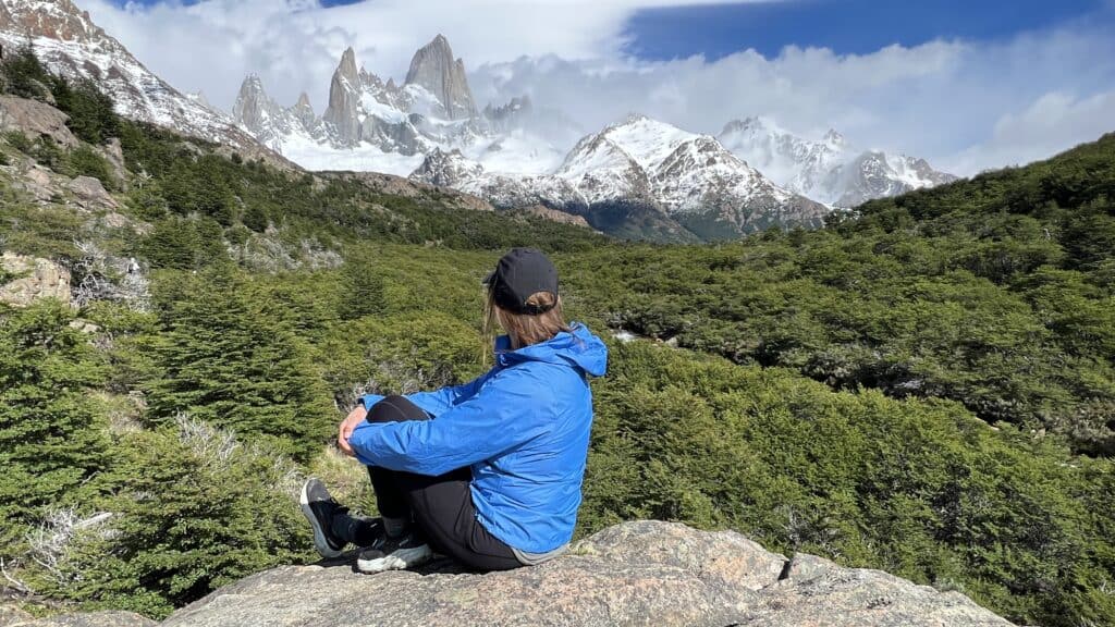 Person in blue jacket sits on a rock, planning a 1 week Patagonia itinerary while gazing at snow-capped mountains and green forest.