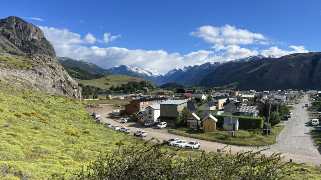 Small town in a green Patagonian valley with parked cars, surrounded by mountains and a partly cloudy blue sky.