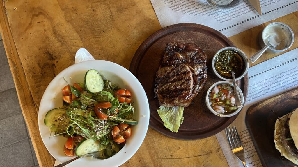 A grilled steak with sauces on a wooden plate, inspired by Patagonia, served next to a fresh salad of cucumbers, tomatoes, and greens.