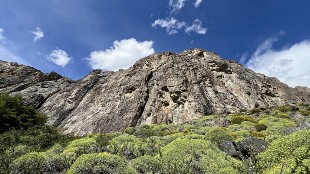 Steep rocky cliff in Patagonia with sparse vegetation at the base, under a bright blue sky with a few clouds.