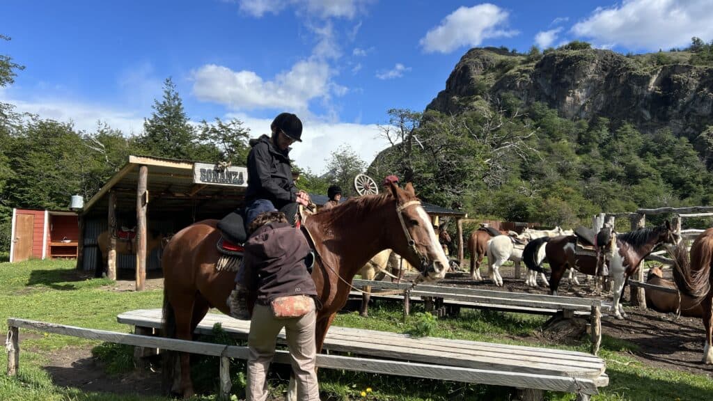A person helps another onto a brown horse at a Patagonia ranch, with rocky hills and more horsesโperfect for your 1 week itinerary.