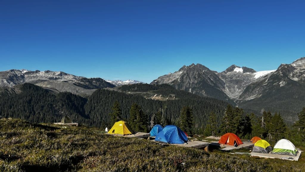 Colorful tents are set up on a grassy hillside, perfect for Pacific Crest Trail hiking with mountains and a clear blue sky in the background.