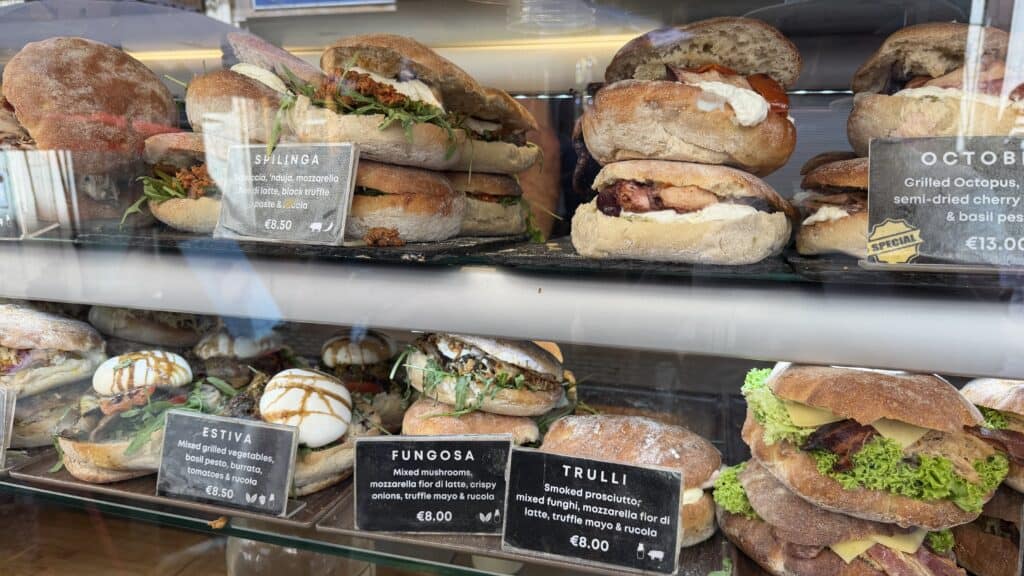 A display case filled with gourmet sandwiches—perfect for fueling your 3 day Malta itinerary—each labeled with ingredients and prices.