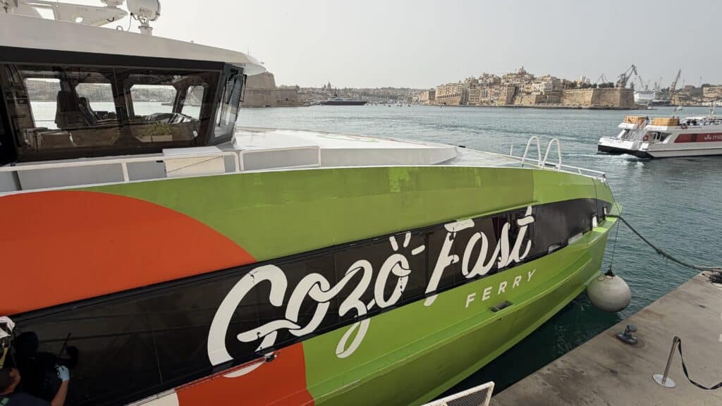 A green and red Gozo Fast Ferry docked at a harbor, perfect for your Malta itinerary, with a city and water taxis in the background.