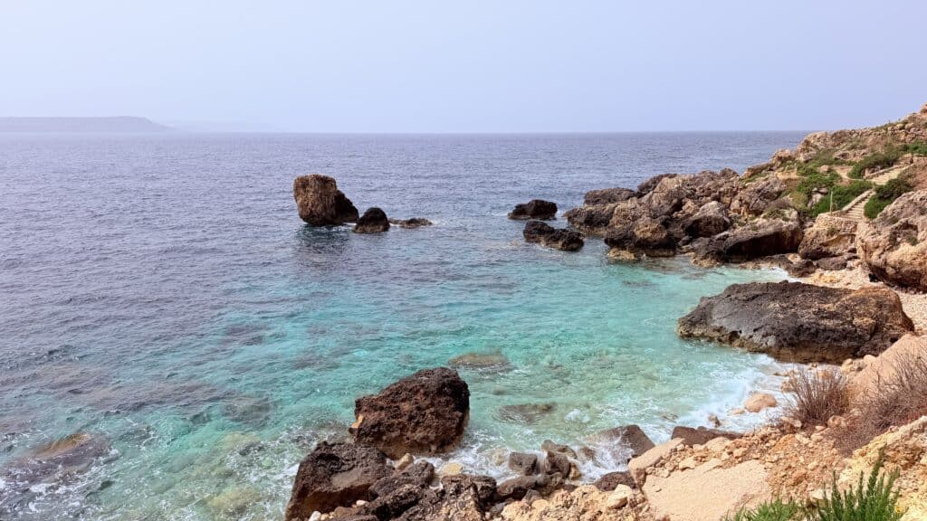 Rocky coastline with clear turquoise water and large rocks, under a hazy sky—ideal for your Malta itinerary or 3 day Malta adventure.