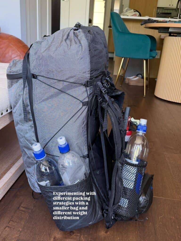 Gray hiking backpack, perfect for Pacific Crest Trail adventures, with mesh pockets and water bottles, on a wooden floor near a dining area.