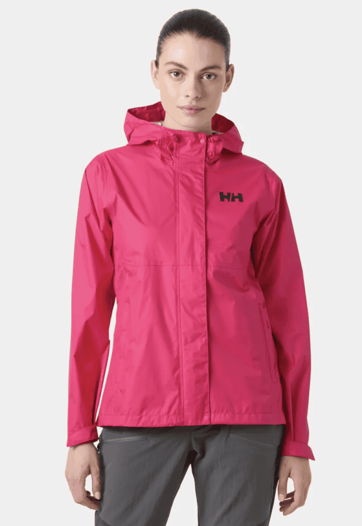 Woman wearing a bright pink hooded Helly Hansen rain jacket, gray pants, and hiking equipment, standing against a plain light background.