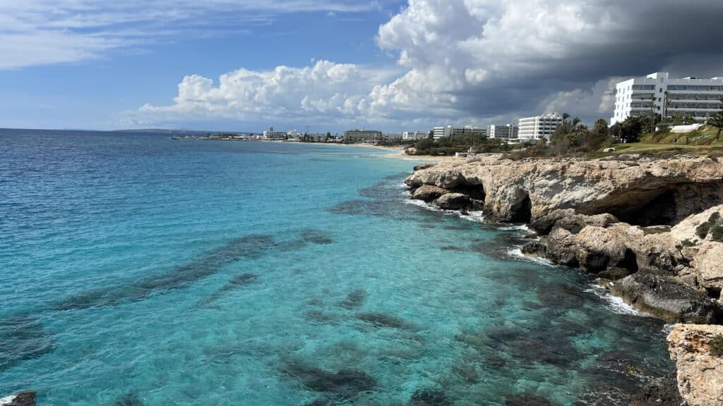 Clear turquoise water meets a rocky shoreline perfect for a 3 day Cyprus itinerary, with coastal hotels in the distance.