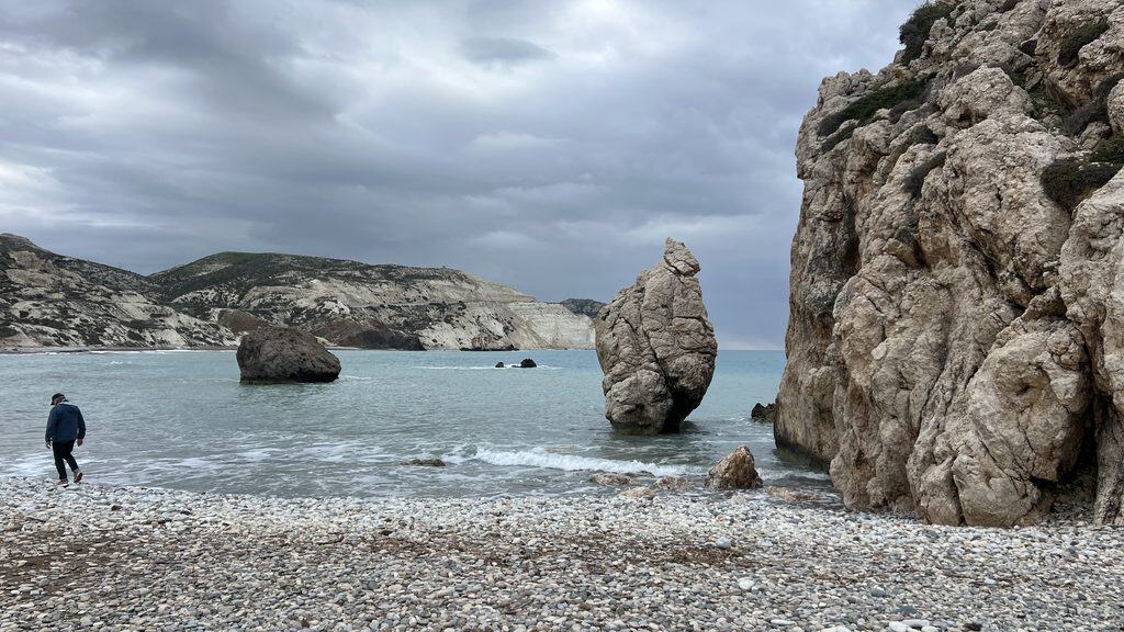 A person stands on a pebble beach facing large rock formations in the sea, a perfect spot for any Cyprus travel guide under a cloudy sky.