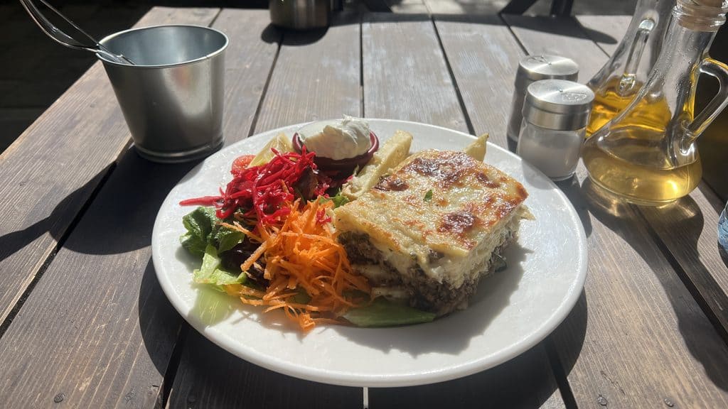 A plate of moussaka, a staple of any Cyprus travel guide, served with shredded carrots, mixed greens, and sour cream on a wooden table.