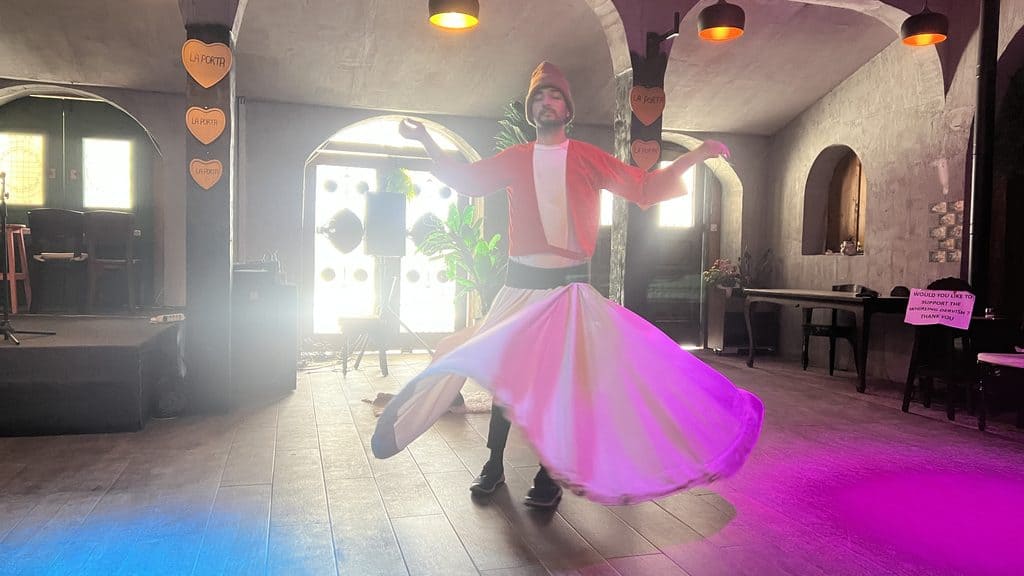 A man in a red jacket and white skirt performs a whirling dervish dance during a 3 day Cyprus trip in a room with arched doorways.