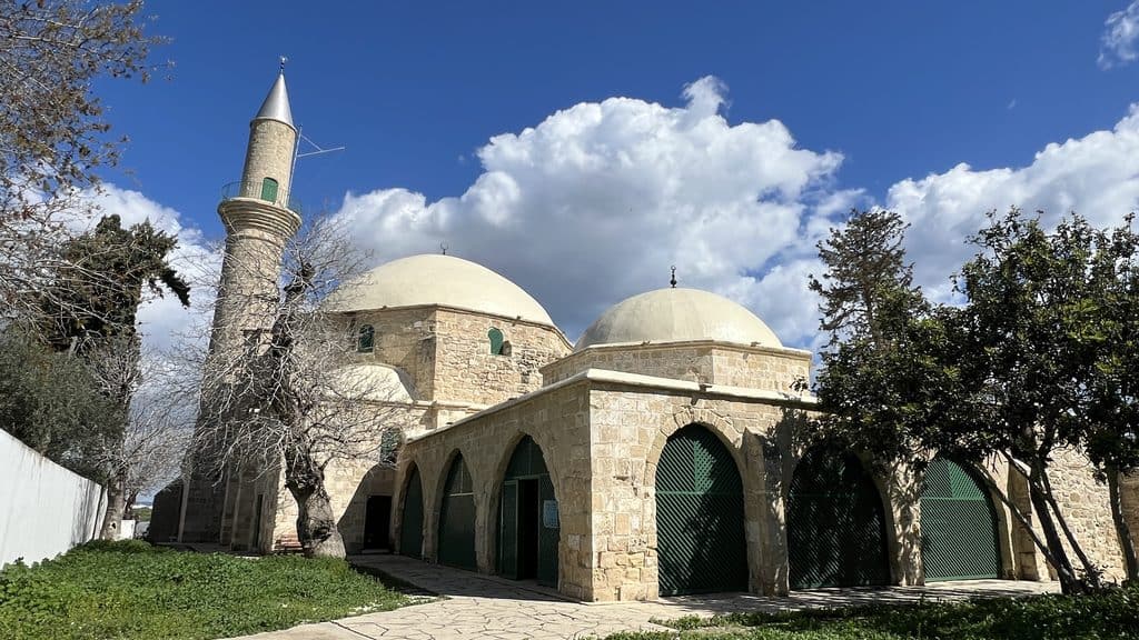 A stone mosque with two domes and a minaret, ideal for a Cyprus itinerary, featuring arched entrances with green doors under a blue sky.